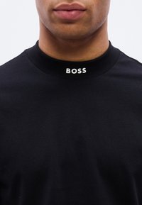 Κοντινό πλάνο ενός ατόμου που φοράει μαύρο μπλουζάκι με στρογγυλ ή λαιμόκοψη, με τη λέξη «BOSS» τυπωμένη σε λευκό στο μπροστινό μέρος του γιακα.