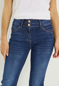 Vaqueros de mezclilla azul oscuro de cintura alta, con dos botones dorados, múltiples bolsillos y un ligero elastano para mayor comodidad.