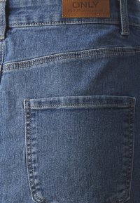 Blaue Jeans aus Denim mit einer Gesäßtasche. Der Bund ist mit einem braunen Lederetikett versehen, auf dem "ONLY" gedruckt ist. Strukturierter, klassischer Stil.