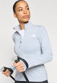 Ανοιχτό γαλάζιο φλις μπουφάν με ψηλό γιακά, φερμουάρ και λογότυπο· διαθέτει οπές για τους αντίχειρες και φοριέται με μπαστούνια πεζοπορίας.