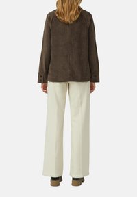 Persoon met schouderlang haar staat met de rug naar ons toe, gekleed in een bruine corduroyjas, witte wijde broek en bruine schoenen op een witte achtergrond.