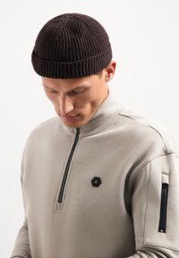 Donkerbruine gebreide muts met een geribbelde rand, gedragen met een lichtgrijze hoodie met ritssluiting, voorzien van een hexagonale badge en een zak met ritssluiting.