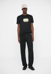 Man in een zwart Armani Exchange T-shirt en broek met gouden logo's, een beige pet en zwarte sneakers, staand voor een witte achtergrond.