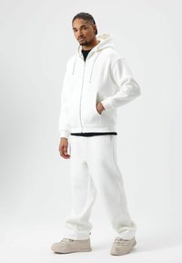 Sweat à capuche en polaire blanche avec cordons de serrage ajustables et poches avant, associé à un pantalon de survêtement blanc assorti, porté par un mannequin.