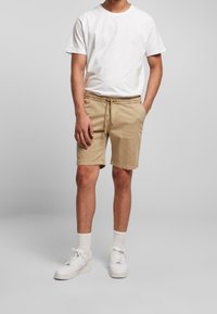 Shorts beiges avec une ceinture élastique et un cordon de serrage, poches latérales, devant plat, associés à un t-shirt blanc uni et des baskets blanches.