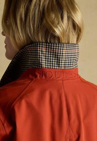 Czerwona wodoodporna kurtka z wysokim kołnierzem w czarno-białą kratę. Na tyle kołnierza tłoczone logo "JOULES". Gładka tekstura.