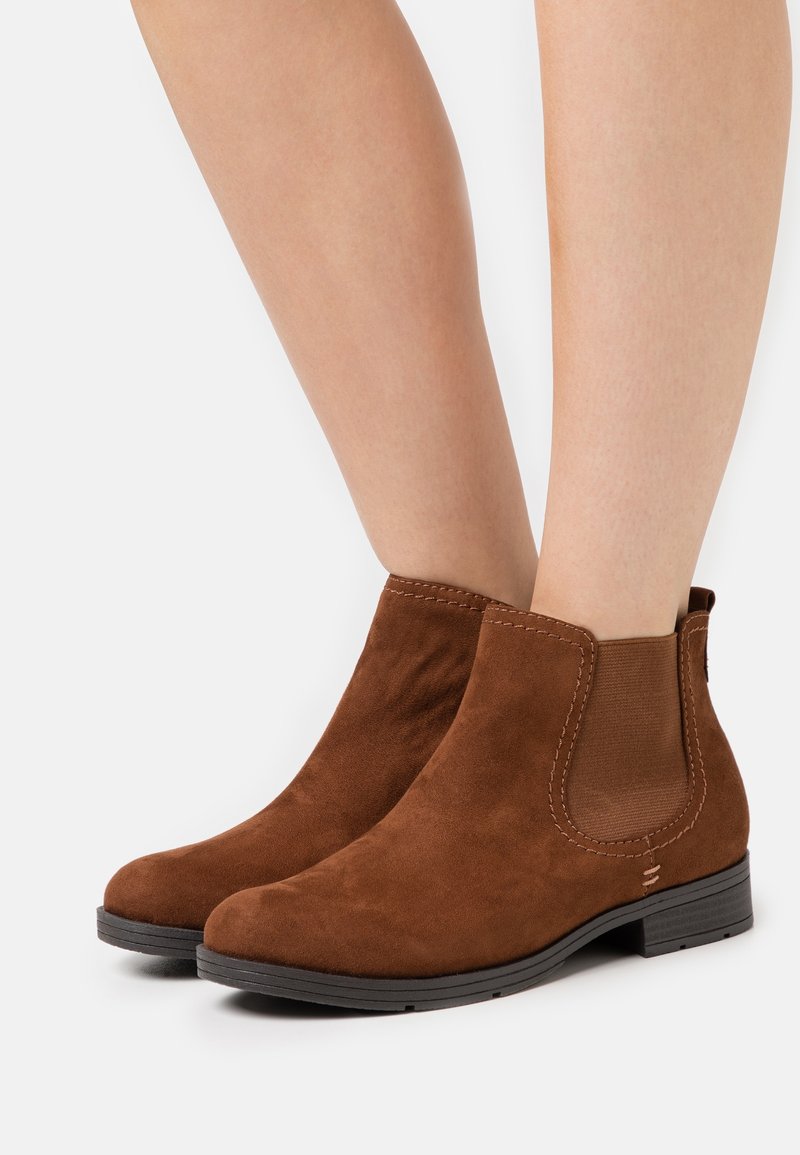 Bottes en suède marron montant à la cheville avec des panneaux latéraux élastiques portées sur des jambes nues, dotées de talons bas noirs et d'orteils arrondis.