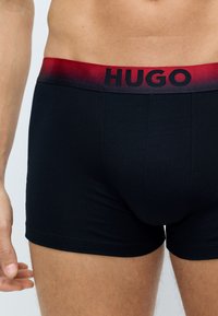 Melna kokvilnas bokseru apakšveļa ar sarkanu gradienta elastīgo jostu, uz kuras ir melnais "HUGO" logo, gluda tekstūra un pieguļošs dizains.