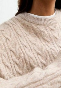 Pull beige en maille torsadée avec un col rond, des poignets côtelés et un tissu doux et texturé. Détails comprenant un motif torsadé en relief.