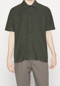Uomo che indossa una camicia a maniche corte verde scuro con bottoni e pantaloni marrone chiaro, in piedi con le braccia rilassate lungo i fianchi su uno sfondo bianco.