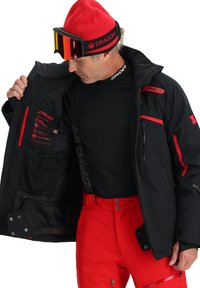 Giacca da sci nera resistente alle intemperie con accenti rossi, tasche con zip e loghi. Sotto, un capo base aderente nero e pantaloni da sci rossi.