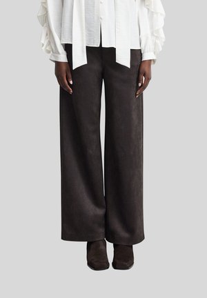 Personne portant un pantalon noir à jambes larges, des chaussures noires et un chemisier blanc à manches longues avec des manches volantées, debout devant un fond clair.