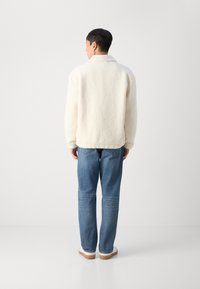 Cremefarvet fleecejakke med blød tekstur, parret med lyseblå jeans og hvide sneakers, set fra ryggen.