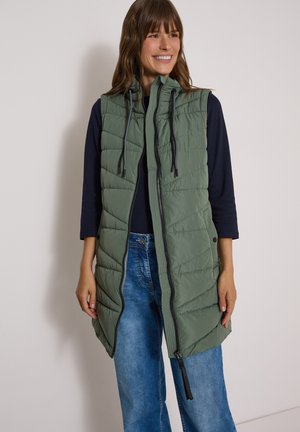 Femme souriante portant une veste matelassée sans manches vert olive par-dessus une chemise noire à manches longues et un jean bleu sur un fond uni.