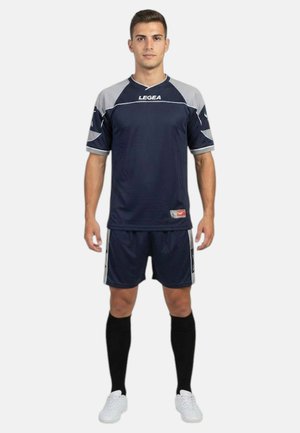 Giovane uomo in piedi che indossa una divisa da calcio blu scuro e grigia con calzettoni neri al ginocchio e scarpe da ginnastica bianche su sfondo bianco.