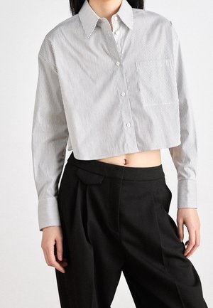 Kobieta ubrana w skróconą koszulę na guziki z długim rękawem, o cienkich szarych paskach, i czarne, plisowane spodnie z wysokim stanem.