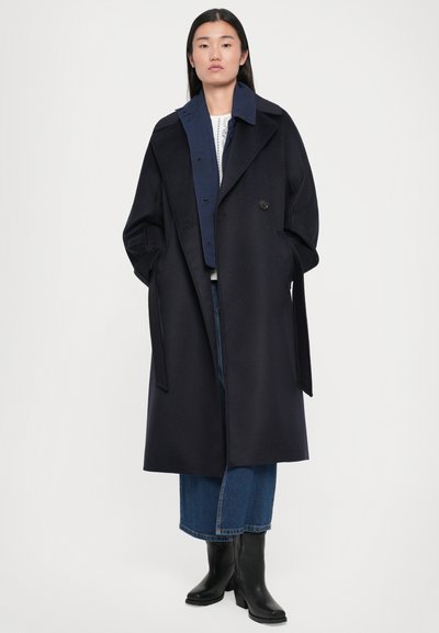 Long navy coat with wide lapels, single button closure, side pockets, and roll-up sleeves over a ribbed white top and denim skirt. Black heeled boots.