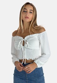 Blouse blanche à épaules dénudées avec des manches longues, accent de nœud à l'avant et détails froncés. Portée avec une jupe en denim bleu.