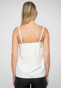 Witte mouwloze top met elastische gerimpelde achterkant, dunne verstelbare zilveren bandjes en een gladde stofstructuur. Toont een eenvoudig ontwerp.
