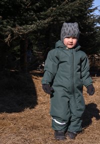 Tuta impermeabile verde intera con cerniera, muffole e striscia riflettente. Accessori includono un cappello grigio con motivo e stivali neri.