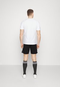 Valkoinen urheilupaita, mustat shortsit ja polven yli ulottuvat sukat, joissa on kaksi valkoista raitaa. Kankaan pinta näyttää sileältä; shortseissa on sivureiät.