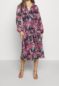 Flödande marinblå klänning med rosa och vita blommönster, V-hals, långa puffärmar, insvängd midja och ankellång fåll. Har ett knytbälte.