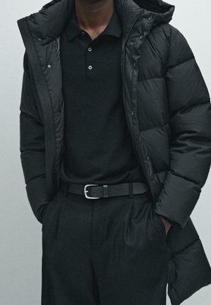 Mann trägt eine schwarze Steppjacke über einem schwarzen Poloshirt, dazu schwarze Hose und schwarzen Gürtel, Hände in den Taschen, vor neutralem Hintergrund.