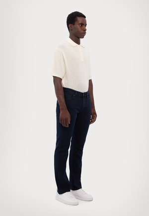 Camisa de polo blanca de punto con cuello, combinada con unos vaqueros azul oscuro y zapatillas blancas, de pie contra un fondo neutral.
