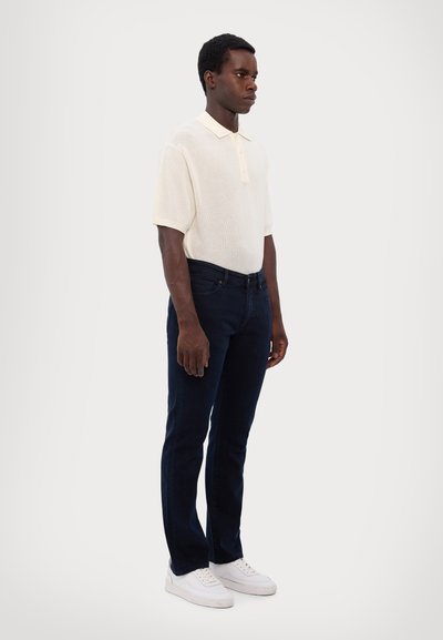 Weißes Strickpolohemd mit Kragen, kombiniert mit dunkelblauen Jeans und weißen Sneakers, vor einem neutralen Hintergrund stehend.