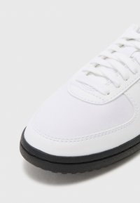Hvide sneakers med et tekstureret stofoverdel, rund tå og traditionelt snørebåndsdesign. Har sort gummisål og kontrasterende syninger.