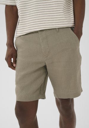 Mand iført beige linnedshorts og en beige og hvid stribet t-shirt, med den ene hånd i lommen, beskåret ved midt på låret.