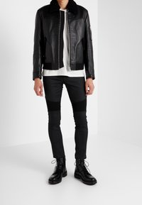 Chaqueta de cuero negra con cuello de borreguito, frente con cremallera, combinada con pantalones negros ajustados con detalles en las rodillas y botas negras con cordones.