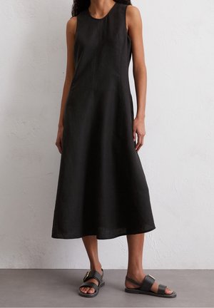 Woman wearing a sleeveless, black midi dress with side seams, paired with black flat sandals with gold buckles, standing on a gray floor.