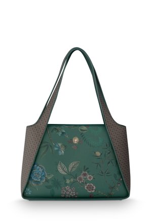 Woman with long dark hair wearing a shiny light green blouse and dark pants stands holding a floral patterned handbag indoors.