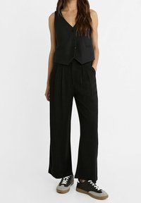 Zwarte mouwloze vest met drie knopen en twee zijzakken, gecombineerd met een brede zwarte broek en zwart/grijze sneakers.