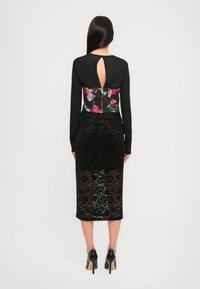 Black long-sleeve dress with floral-patterned bodice and keyhole back detail, featuring a lace overlay skirt and high-heeled shoes.