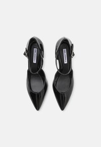 Crne lakirane stiletto cipele s šiljastim vrhom, remenom oko članka i detaljem od srebrne kopče. Glatka tekstura s sjajnim završetkom.