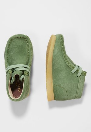 Zielone zamszowe buty z zaokrąglonym noskiem, sznurowane, z jasnobrązową gumową podeszwą. Charakteryzują się widocznym przeszyciem i miękką teksturą.