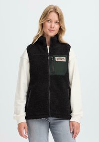 Vrouw in een zwart fleece zip-up vest met een borstzaklabel "Ontworpen met Toewijding" over een witte sweatshirt en lichtblauwe jeans.