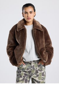 Ruskea tekoturkistakki leveällä kauluksella, vetoketjulla ja rennolla istuvuudella, yhdistettynä maastokuvioisiin housuihin, joissa on abstrakteja muotoja.