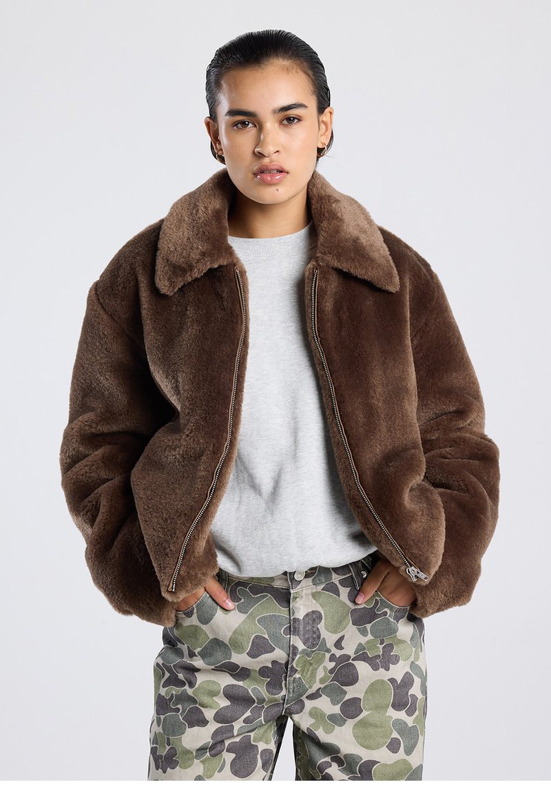 Ruskea tekoturkistakki leveällä kauluksella, vetoketjulla ja rennolla istuvuudella, yhdistettynä maastokuvioisiin housuihin, joissa on abstrakteja muotoja.