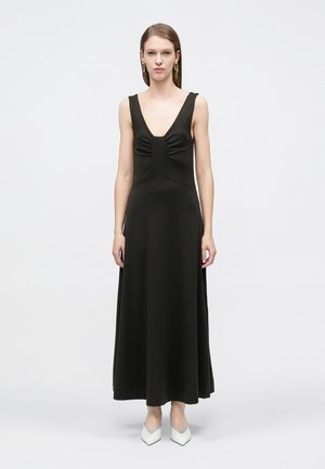 Kobieta ubrana w czarną, bezrękawnikową suknię maxi z głębokim dekoltem w kształcie litery V oraz białe szpilki z ostrym noskiem, stojąca na jednolitym tle.