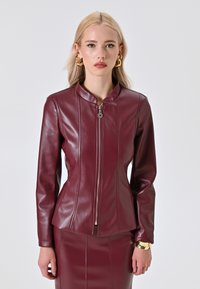Veste en cuir synthétique bordeaux avec une coupe ajustée, col haut, motif matelassé et fermeture éclair argentée. S'associe avec une jupe moulante.