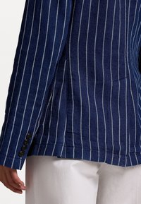 Navy striped linen jacket featuring vertical white stripes, three black buttons on the cuff, and a tailored fit with a side slit detail.