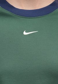 Πράσινο πουλόβερ με λαιμόκοψη και ναυτική γιακά και λευκό λογότυπο Nike Swoosh κεντραρισμένο στο στήθος κάτω από το λαιμό.