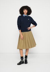 Tengerészkék pulóver bordázott mandzsettával, farmeringgel rétegezve; pliszírozott bézs szoknya; fekete cipők fekete-fehér csíkos zoknival.