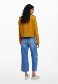Senapsgul läderjacka med kort skärning, detail på ryggen och axelremmar; kombinerad med ljusblå vida jeans.