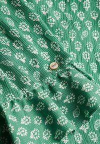 Tejido verde con un patrón texturizado de motivos florales blancos. Se puede ver una etiqueta metálica redonda con detalles de marca, que añade un acento de diseño.