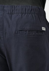 Navy fabric pants with an elastic waistband, featuring a back pocket and a small brand label stitched on the side. Smooth texture.