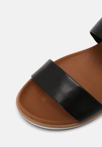 Ruskea sandaalin pohja, jossa on leveä musta nahkahihna edessä ja nilkkahihna, joka näkyy valkoista taustaa vasten.